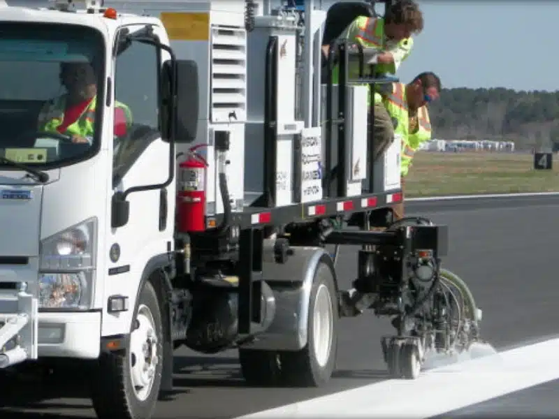 Pavement marking crew operating striping truck to apply white road lines during highway construction project.