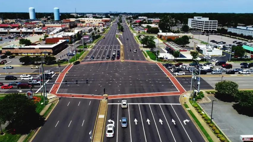 Aerial view of a wide urban intersection with multiple traffic lanes, turn arrows, and crosswalks surrounded by commercial buildings.